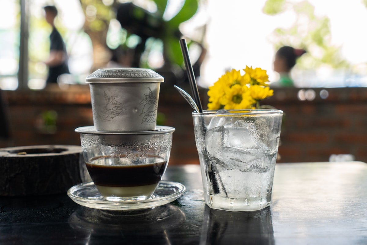 A Vietnamese phin filter brewing coffee next to a glass of ice with yellow flowers